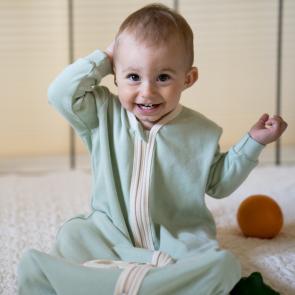 Baby sitting up and smiling at the camera. Baby is wearing a long sleeve mint green sleeper bag with natural trim and zipper in the center front. 